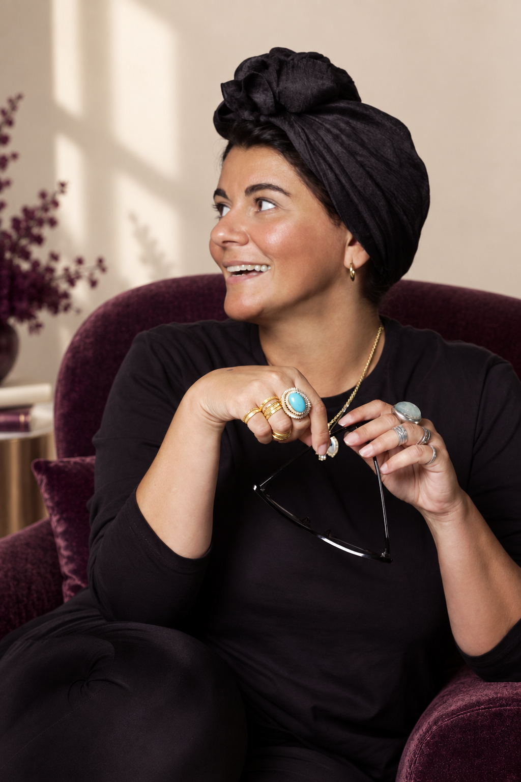 Asil Attar seated in a plum velvet chair, wearing a black turban and jewellery, holding glasses