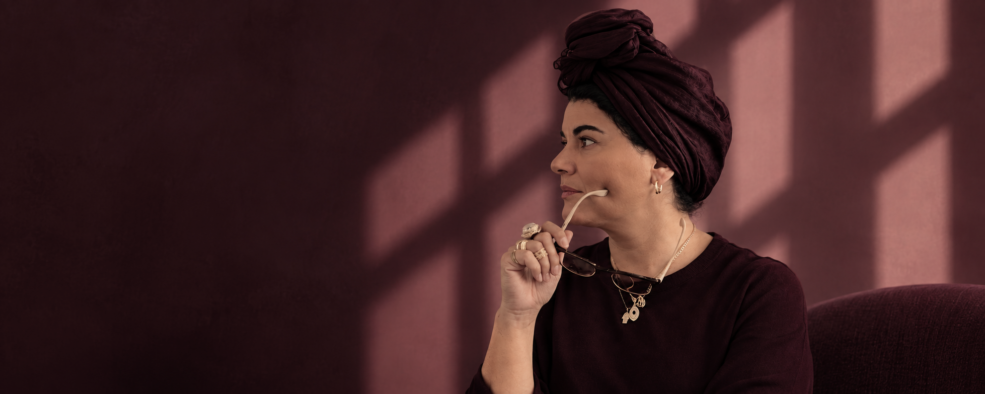 Asil Attar in a plum turban holding her glasses, seated against a plum wall with soft window-light shadows